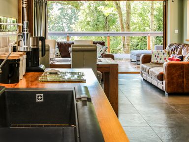 A kitchen sink with countertop and coffee machine next to a dining table and brown leather sofa in a living area with glass door opening to a balcony with trees outside at Sunnyside in Bircher Common near Yarpole