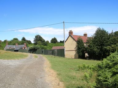 A dirt road leading past houses with fences and trees under a blue sky at Sunnyside in Bircher Common near Yarpole