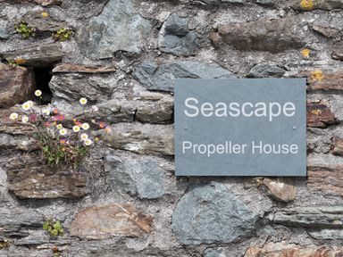 A stone wall with a sign reading Seascape Propeller House and flowers growing in the crevices