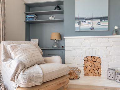 An armchair with a cushion and throw in front of a white brick fireplace with stacked firewood and built-in shelves with books and a lamp at Seascape in Salcombe
