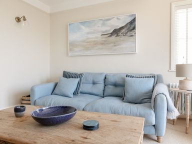 A blue sofa with cushions and a blanket in a living room with a wooden coffee table and a landscape painting on the wall at Seascape in Salcombe