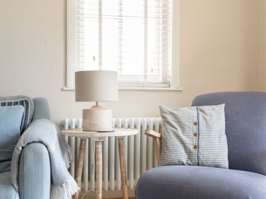 A wooden side table with a lamp between a blue sofa with a blanket and a blue chair with a striped pillow at Seascape in Salcombe