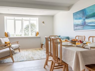 A dining area with a table set for breakfast wooden chairs a seating area with armchairs and cushions by a window at Seascape Salcombe