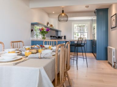 A dining table set with plates orange juice and bread in a kitchen with blue cabinets and wooden chairs at Seascape Salcombe