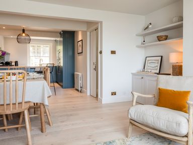 A dining area with wooden chairs and table next to a kitchen with blue cabinets and a lounge chair with an orange pillow at Seascape in Salcombe