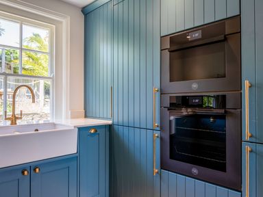 A kitchen corner with blue cabinetry white countertop a farmhouse sink with a brass faucet and built in black ovens at Seascape in Salcombe