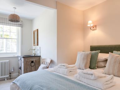 A bedroom with a bed with folded towels pillows and a blue throw a wooden chair a rustic sink cabinet and a window at Seascape in Salcombe