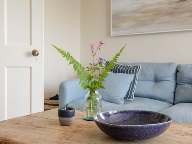 A living room with a wooden table holding a purple bowl a candle and a vase with flowers in front of a blue sofa at Seascape in Salcombe