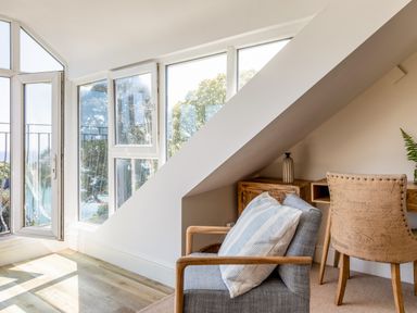 A room with a wooden framed armchair with cushions wooden desk and chair and open windows showing trees at Seascape in Salcombe