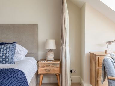 A bedroom with a bed, bedside table with a lamp, curtain, wooden dresser and a decorative bird at Seascape Salcombe