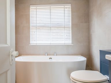 A bathroom with a white bathtub below a window with blinds and a toilet beside a dark cabinet at Seascape in Salcombe