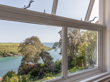 A window view overlooking trees and a body of water with a coastline in the distance at Seascape in Salcombe