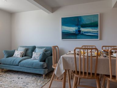 A blue sofa with cushions beside a dining table set with plates and glasses and wooden chairs with a blue abstract painting on the wall at Seascape Salcombe