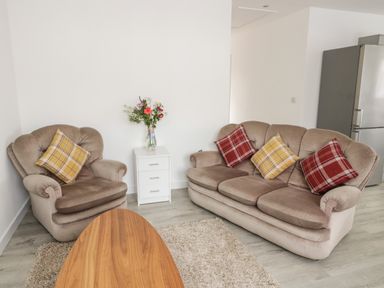 A living room with a beige armchair and sofa with plaid cushions a white drawer unit with flowers and a wooden coffee table at Trigfa in Llanfechell