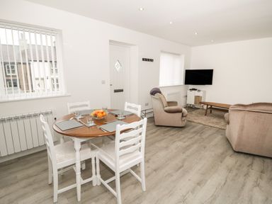 A dining area with a round wooden table set with placemats and glasses next to a living room with two armchairs a wall-mounted TV and a coffee table at Trigfa in Llanfechell