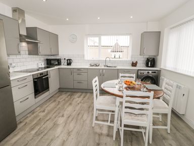 A kitchen with grey cabinets a round wooden table with four white chairs and kitchen appliances at Trigfa in Llanfechell