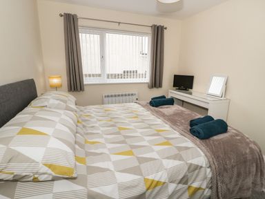 A bedroom with a bed a bedside lamp a window with curtains and a dressing table with a mirror and a TV at Trigfa in Llanfechell