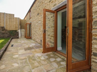 A stone exterior wall with wooden framed glass doors opening onto a paved patio next to a small grass area at The Granary Burton Bradstock
