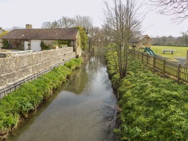 A narrow river with grassy banks and trees next to a stone building and a fenced playground at The Granary Burton Bradstock
