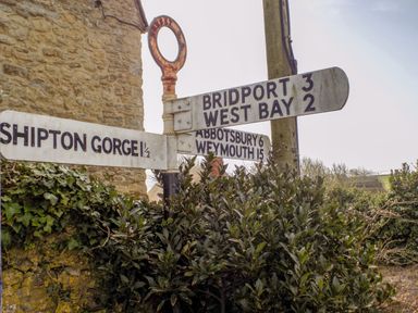A road sign with destinations bridport west bay shipton gorge abbotsbury and weymouth near a stone building with bushes at The Granary Burton Bradstock