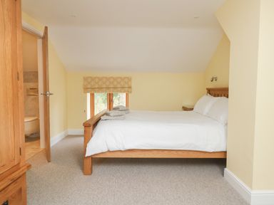 A bedroom with a wooden bed, white bedding, folded towels on the bed, a wooden wardrobe, an open door to a bathroom, and a window with a patterned blind at The Granary Burton Bradstock