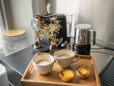A wooden tray with two cups of coffee two pastries and a small vase with dried flowers on a kitchen counter