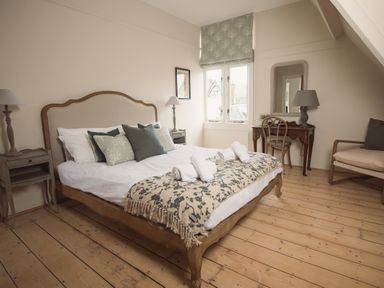 A bedroom with a wooden bed with pillows and rolled towels bedside tables with lamps a window with a floral blind a wooden desk with a chair and a mirror and an armchair at Fairhaven in Dartmouth