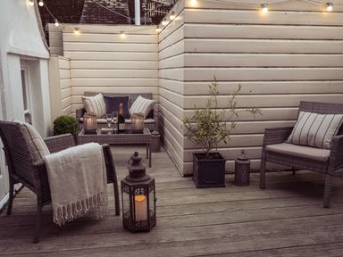 An outdoor seating area with wicker furniture cushions a blanket lanterns a potted plant and string lights on a wooden deck at Fairhaven in Dartmouth