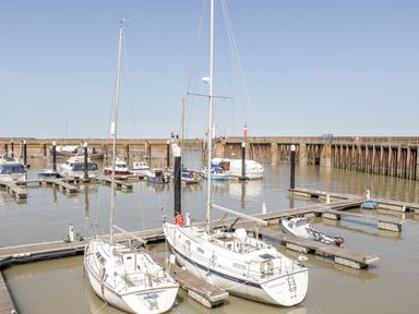 A marina with multiple boats and yachts docked at piers with a concrete wall and lighthouse in the background