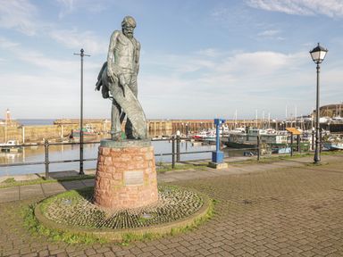 A statue of a man holding a large fish near a harbor with boats and lampposts at The Retreat in Washford near Williton
