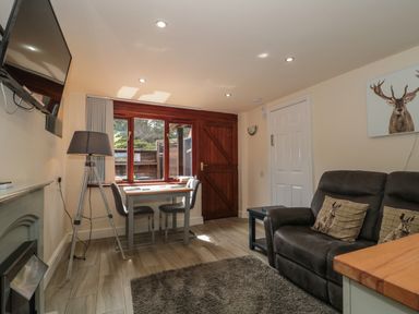 A living room with a dark sofa with animal pillows a small dining table with two chairs a floor lamp a wall-mounted television and a window with a wooden door at The Retreat in Washford near Williton