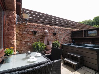 An outdoor patio with a glass table and two wicker chairs two teacups and a teapot a stone water fountain two green plants a hot tub and a wooden fence at The Retreat Washford near Williton