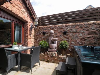 An outdoor patio with a glass table and wicker chairs a stone wall with plants a modern water feature and a hot tub at The Retreat in Washford near Williton