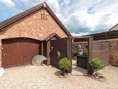 The exterior of a stone building with a wooden double door and a fenced patio area with plants and a sculpture at The Retreat Washford near Williton