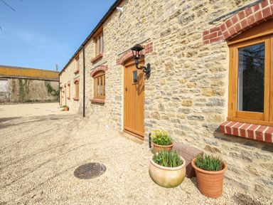 Exterior of a stone house with wooden doors and windows and flower pots on gravel at The Stables Burton Bradstock