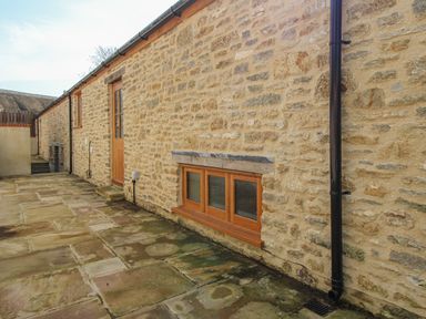 An exterior stone wall with wooden framed windows a wooden door a black drainpipe and a stone paved patio at The Stables Burton Bradstock