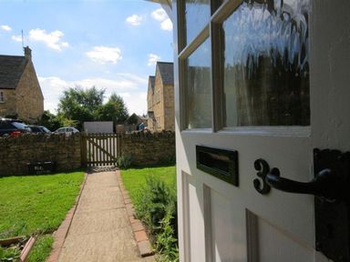A pathway leading from a partially open door to a wooden gate and stone wall with houses and cars in the background at Noel Cottage in Chipping Campden