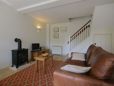 A living room with a brown leather sofa a wooden coffee table a black stove and a wooden media cabinet with a TV at Noel Cottage in Chipping Campden