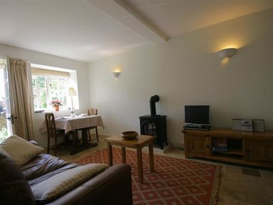 A living room with a brown leather sofa, wooden table, dining table by a window, wood stove, TV on wooden cabinet at Noel Cottage in Chipping Campden