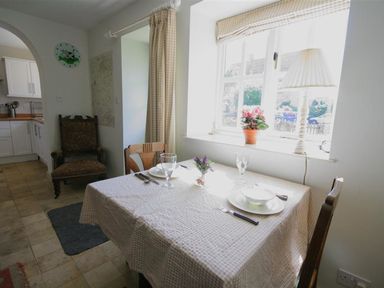A dining area with a table set for two near a window with a lamp and flowers at Noel Cottage in Chipping Campden