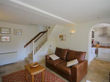 A living room with a brown sofa and wooden table near stairs and an archway to a kitchen at Noel Cottage in Chipping Campden