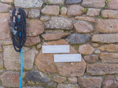 A hose and signs labeled 'SANDY FEET' and 'MUDDY PAWS' on a stone wall at Scilly Stack in Penzance