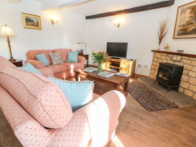 A living room with pink sofas and blue cushions a wooden coffee table with flowers a TV on a wooden stand and a stone fireplace at Cowfair Cottage in Chipping Campden