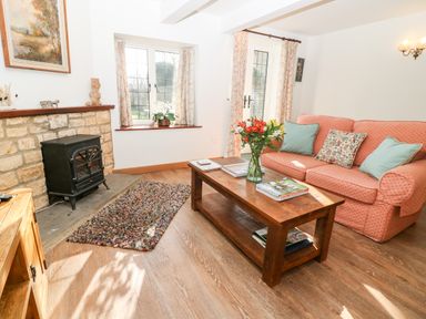A living room with a pink sofa a wooden coffee table flowers on the table a stone fireplace and windows with floral curtains at Cowfair Cottage in Chipping Campden