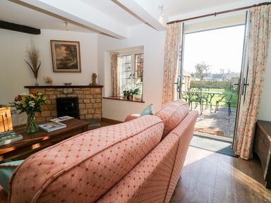 A living room with a patterned sofa a wooden coffee table with flowers an open door leading to a garden and a small stone fireplace at Cowfair Cottage in Chipping Campden