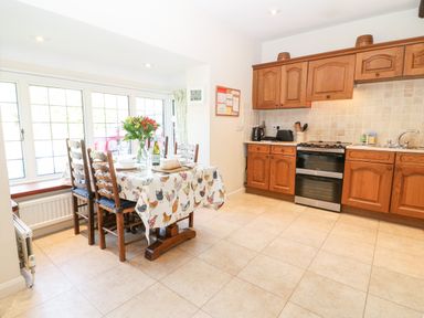A kitchen with wooden cabinets a gas stove a sink and a dining table with six chairs and a floral tablecloth at Cowfair Cottage in Chipping Campden