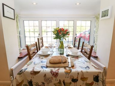A dining area with a table set with bowls glasses a loaf of bread and a bottle with flowers on the table in front of large windows at Cowfair Cottage in Chipping Campden