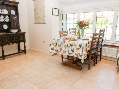 A dining room with a table covered by a chicken patterned tablecloth set with bread flowers and wine near large windows at Cowfair Cottage in Chipping Campden