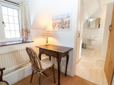 A small wooden desk and chair next to a window with patterned curtains and a lamp on the desk with a view into a bathroom at Cowfair Cottage in Chipping Campden