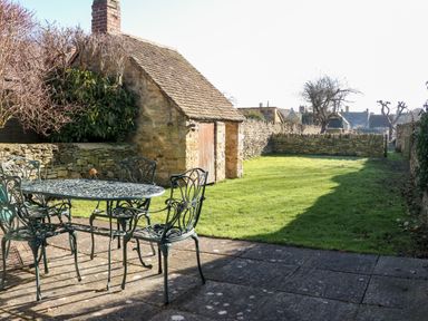 An outdoor patio with metal table and chairs next to a stone shed and a grass lawn at Cowfair Cottage in Chipping Campden